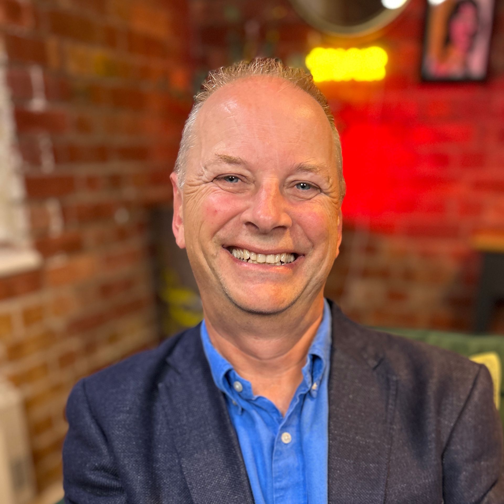 Smiling man in blue shirt and blazer, in front of a brick wall.
