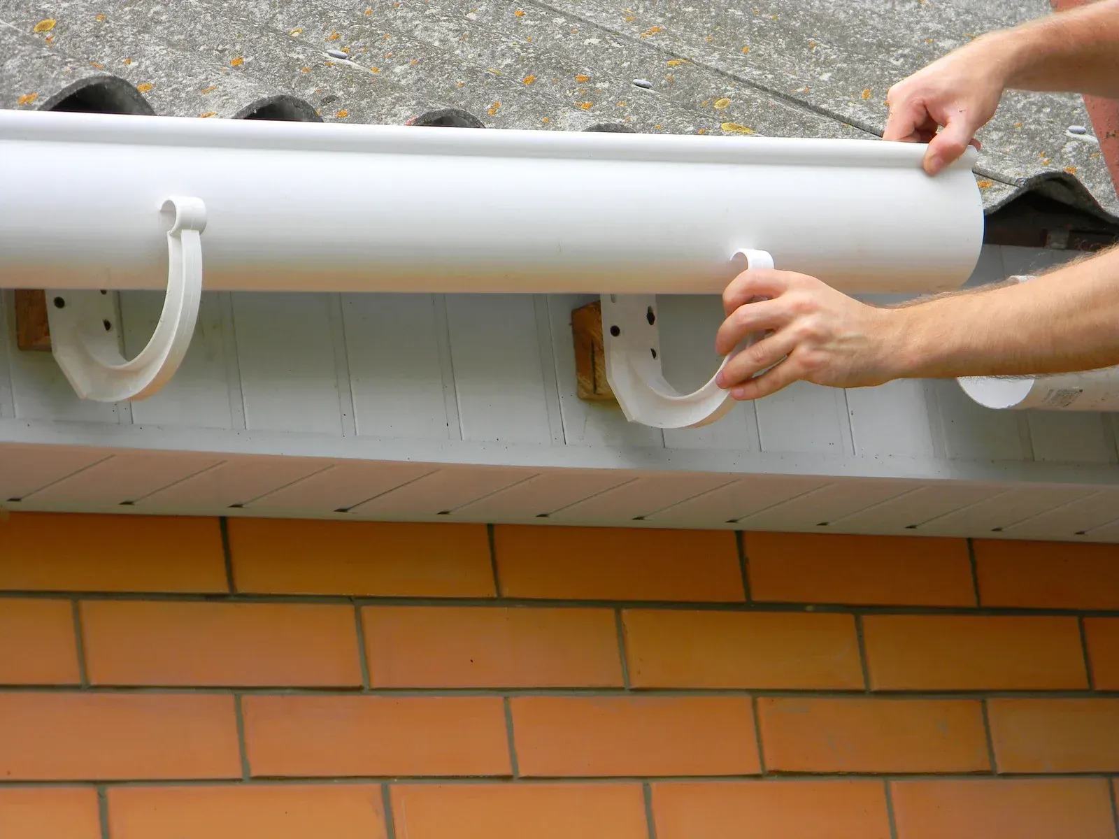 A person is installing a gutter on the side of a brick building.