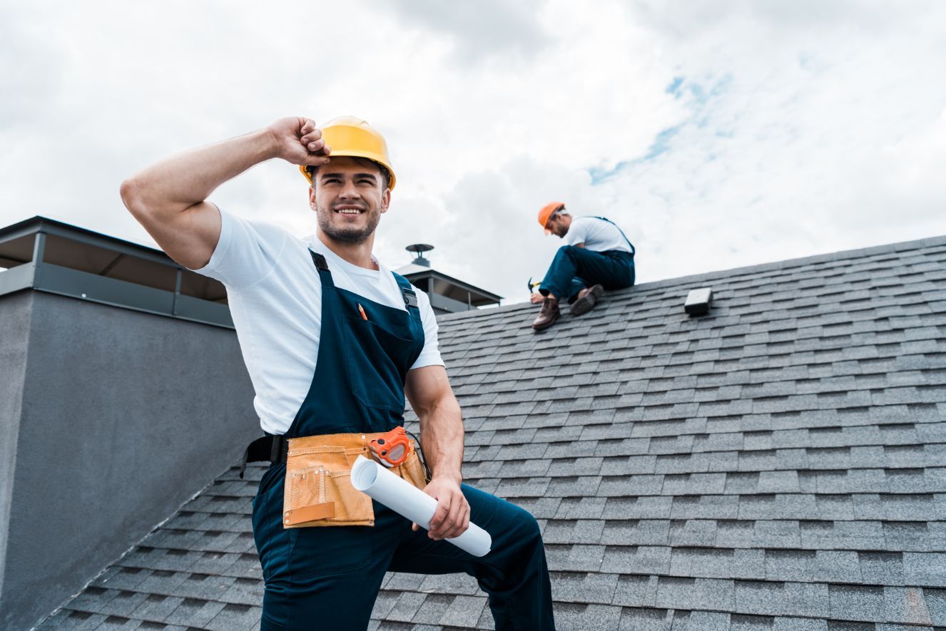 Roofer on a roof, in overalls and hard hat, holds plans, smiles. Another roofer works in the background. Cloudy sky.