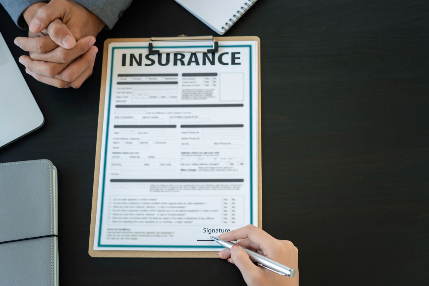 Person signing an insurance document on a clipboard. Another person’s hands visible, along with a laptop and notebook.
