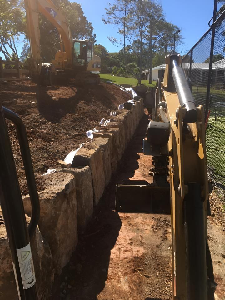A Bulldozer is Digging a Hole in the Ground Next to a Stone Wall — Limited Access Excavation In Murwillumbah, NSW