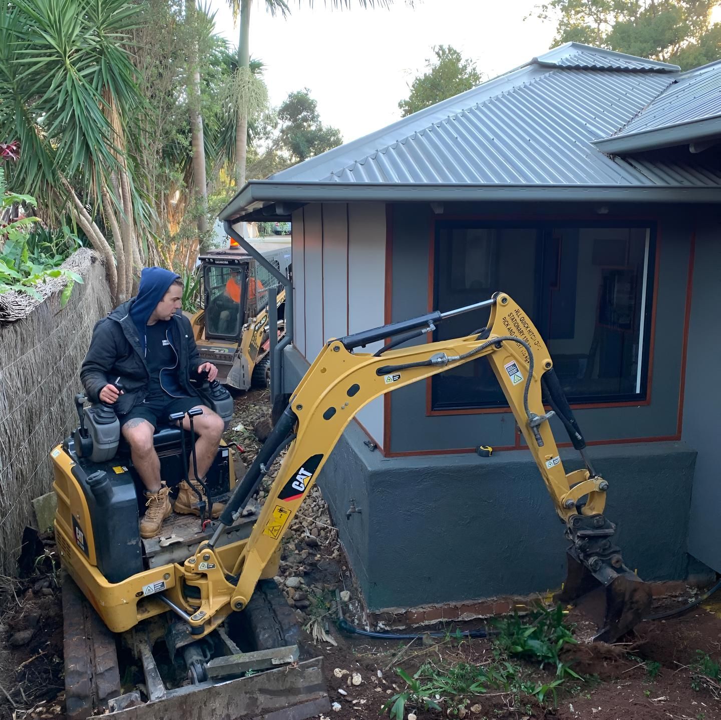 A Man is Sitting on an Excavator in Front of a House — Limited Access Excavation In Lismore, NSW