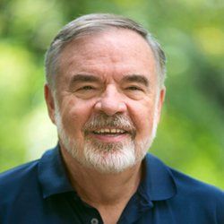 Smiling man with a white beard wearing a blue shirt, outside with blurred green background.