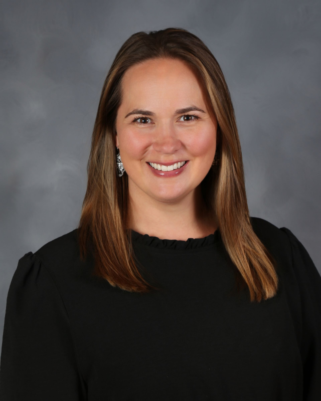 Woman with brown hair smiles, wearing a black top against a grey background.