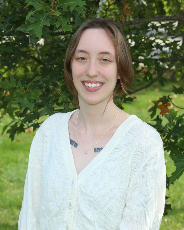 Woman with light brown hair, smiling, wearing a white sweater, standing outside with green foliage in the background.