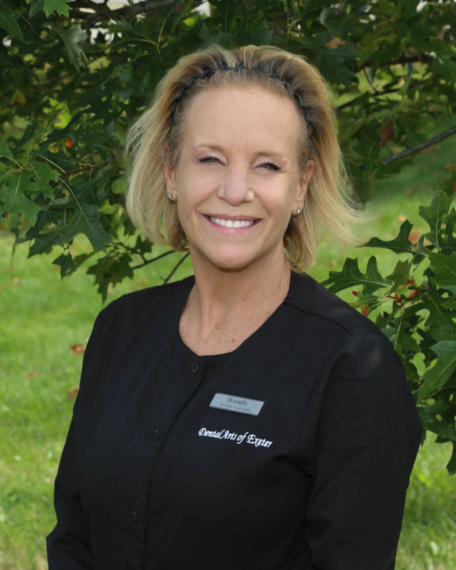 Woman in a black top smiles, standing outdoors near green foliage.