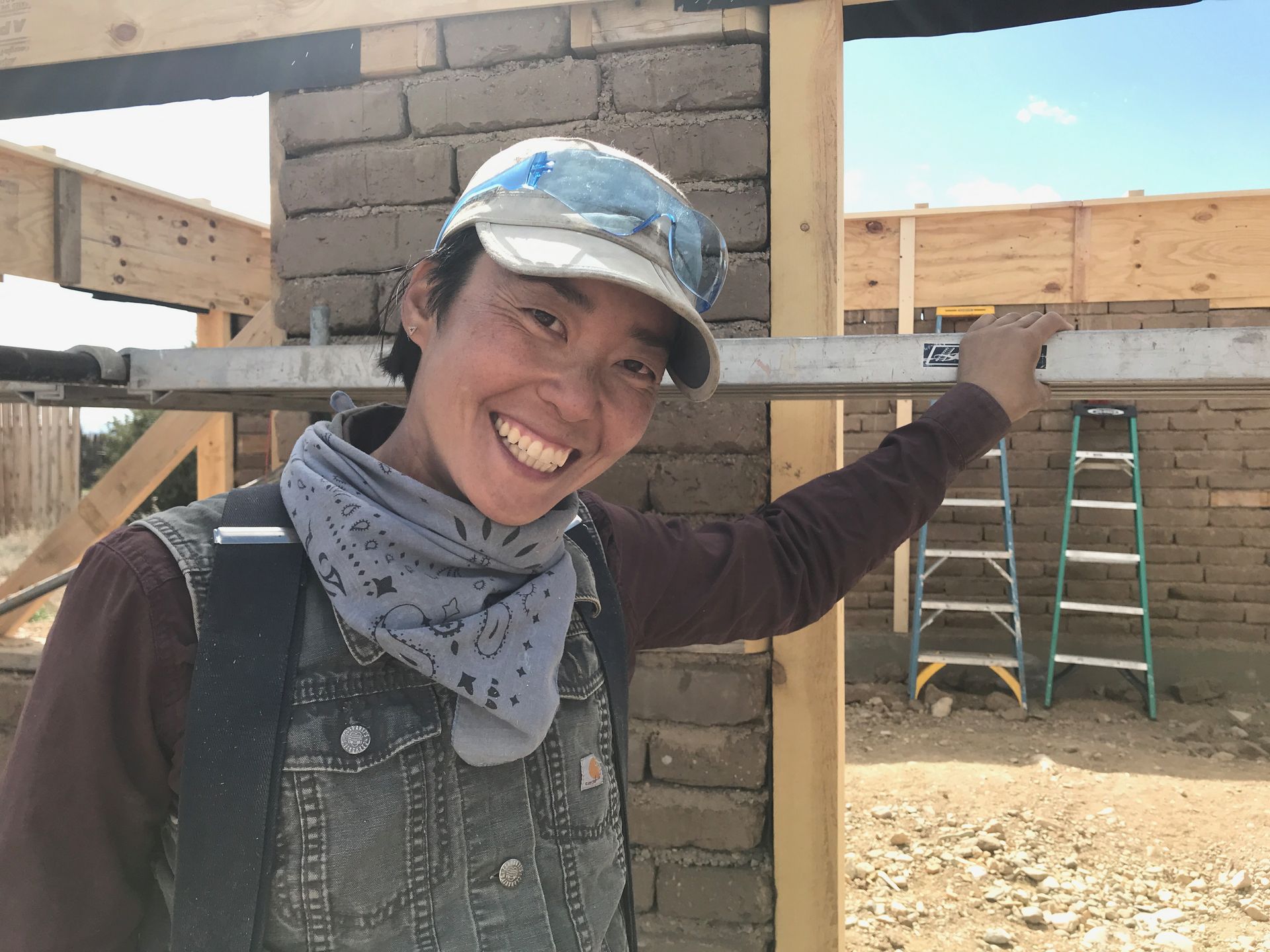 Smiling person in a denim vest building a mud-brick wall on a sunny day. Construction materials and a ladder are visible in the background.