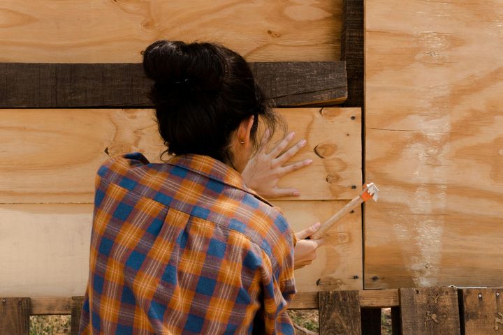 A woman in a plaid shirt hammers wood planks onto a wooden structure outdoors.