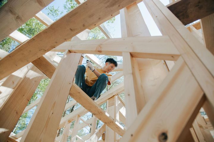 A person building wooden framework for a house, blue jeans and yellow shirt. Sunlight streams through the wooden structure.
