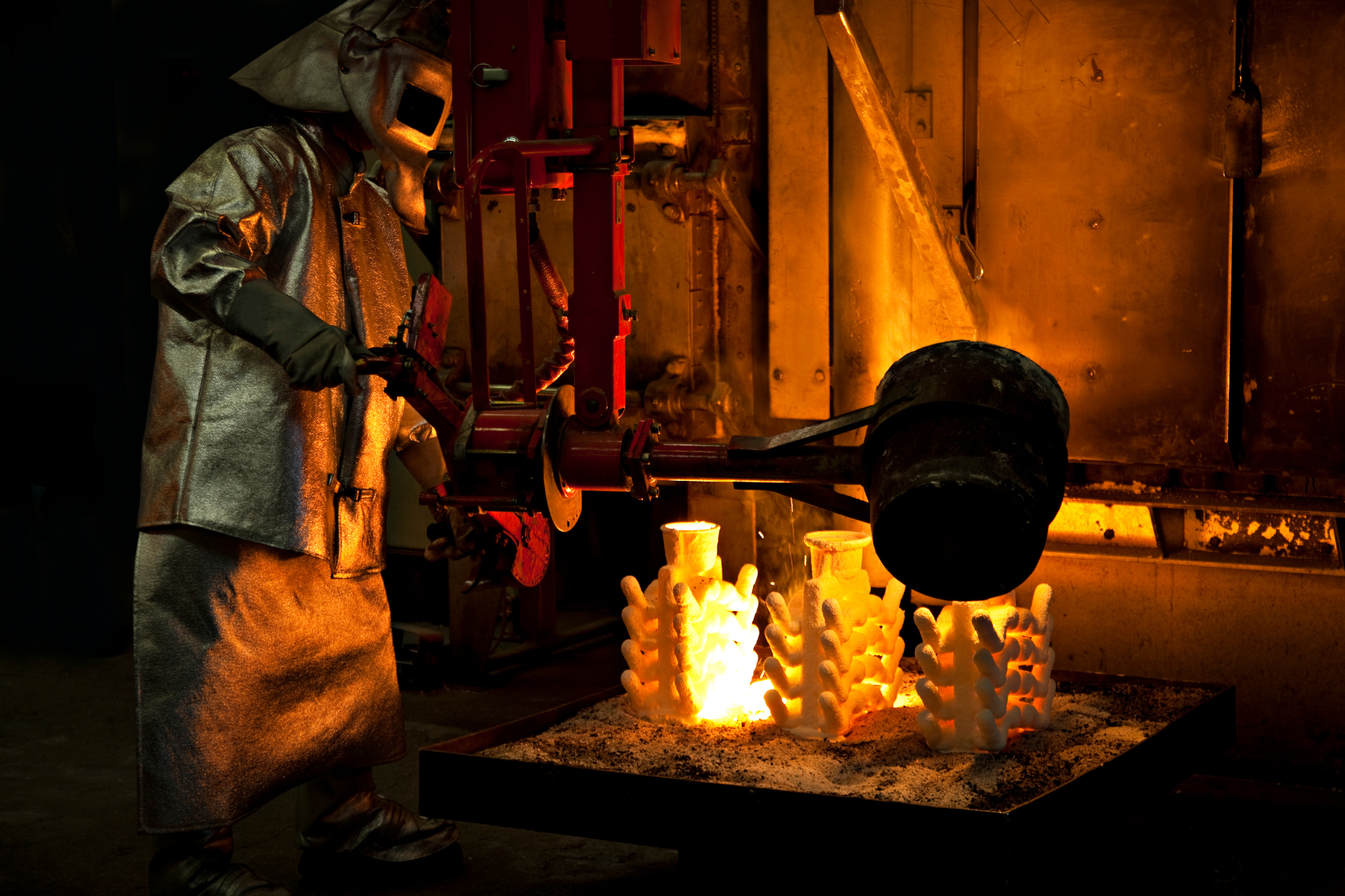 A worker in protective gear pours molten metal onto molds in a foundry, with an orange-lit interior.