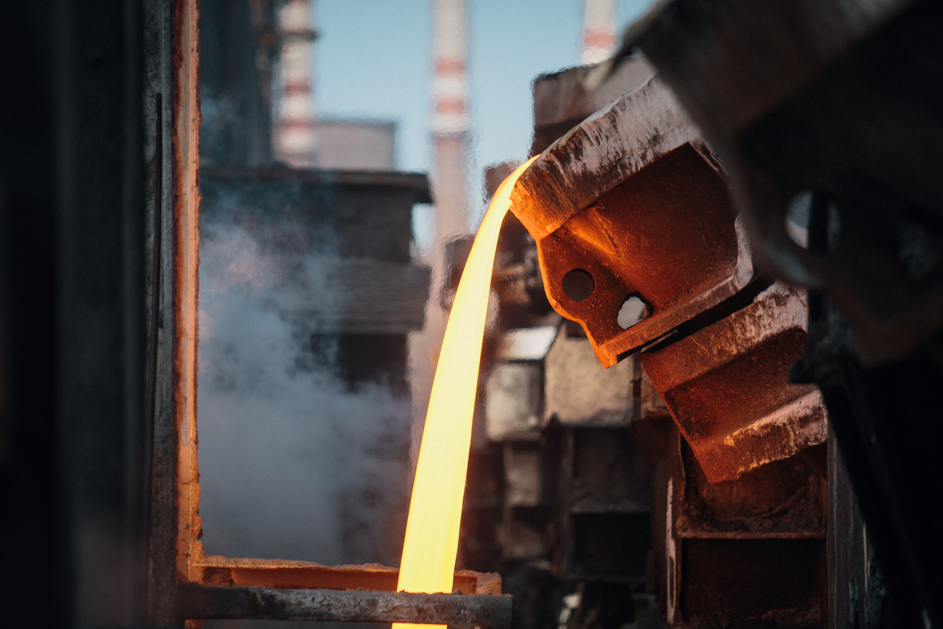 Molten metal pouring from a furnace, orange glow. Factory setting, smoke rising.