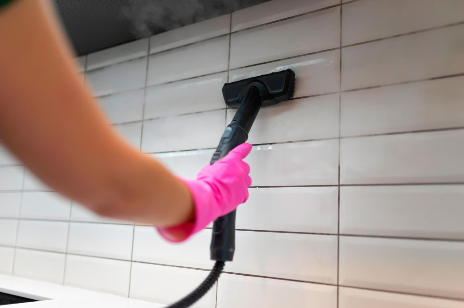 Una persona que lleva guantes rosas está limpiando una pared de azulejos con un limpiador a vapor.