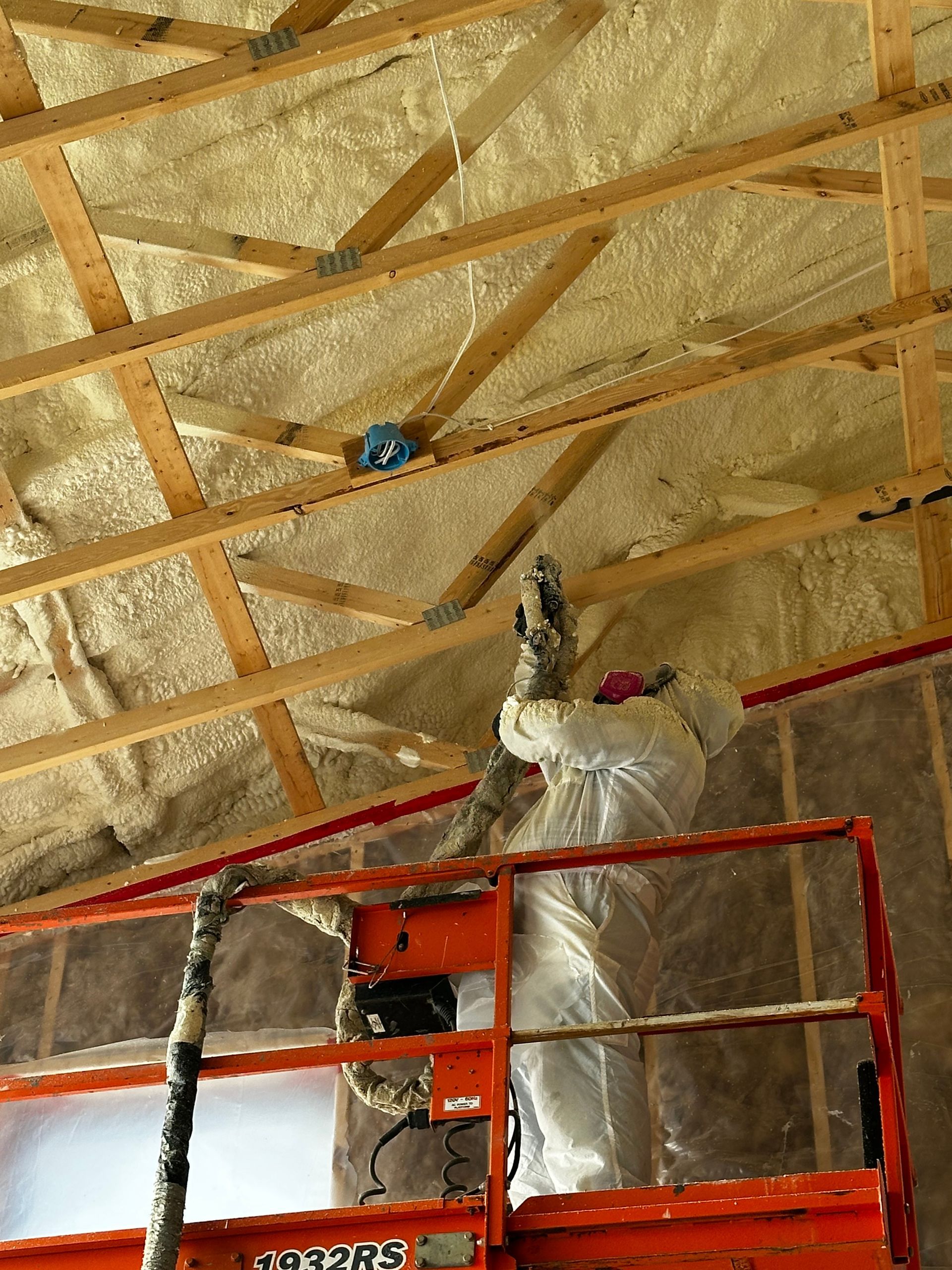 A worker in protective gear on a lift sprays foam insulation onto the wooden rafters of a building ceiling.