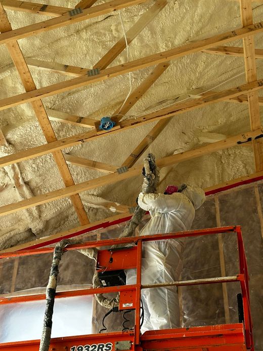 A worker in protective gear on a lift sprays foam insulation onto the wooden rafters of a building ceiling.