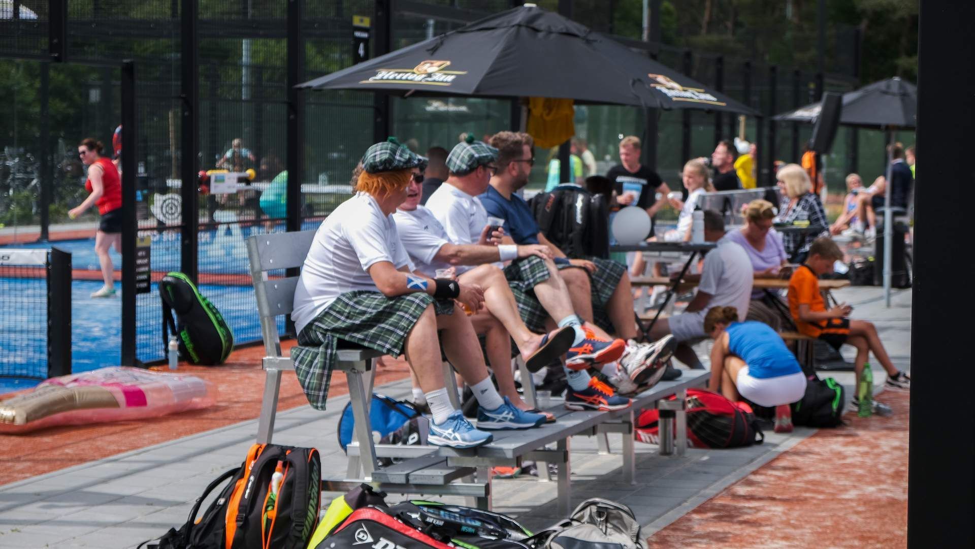 Mensen zitten op bankjes naast een padelbaan onder parasols en kijken naar de spelers tijdens een evenement in de openlucht.
