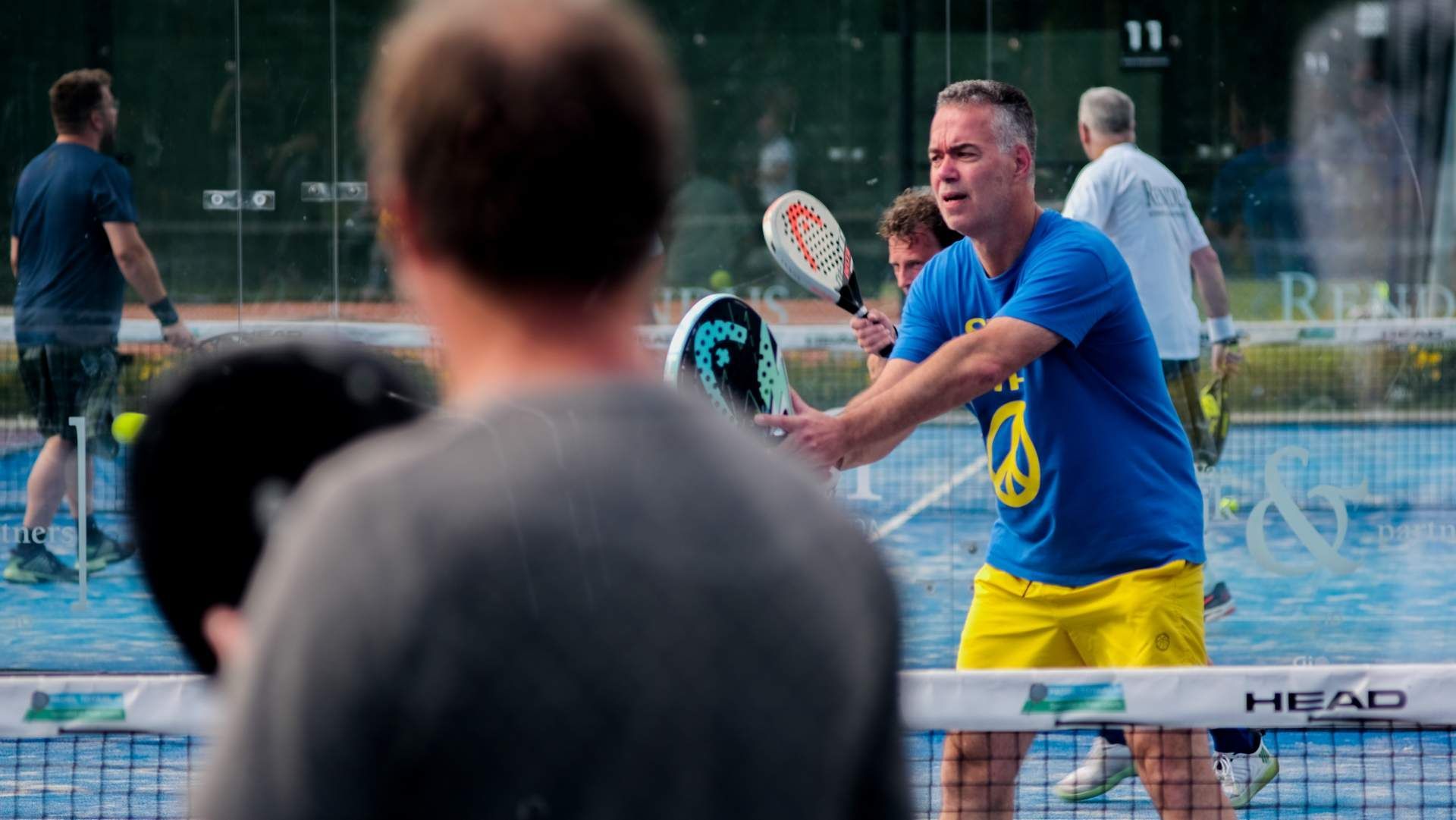 Mensen spelen padel op een buitenbaan, met op de voorgrond een speler in een blauw shirt en een gele korte broek.