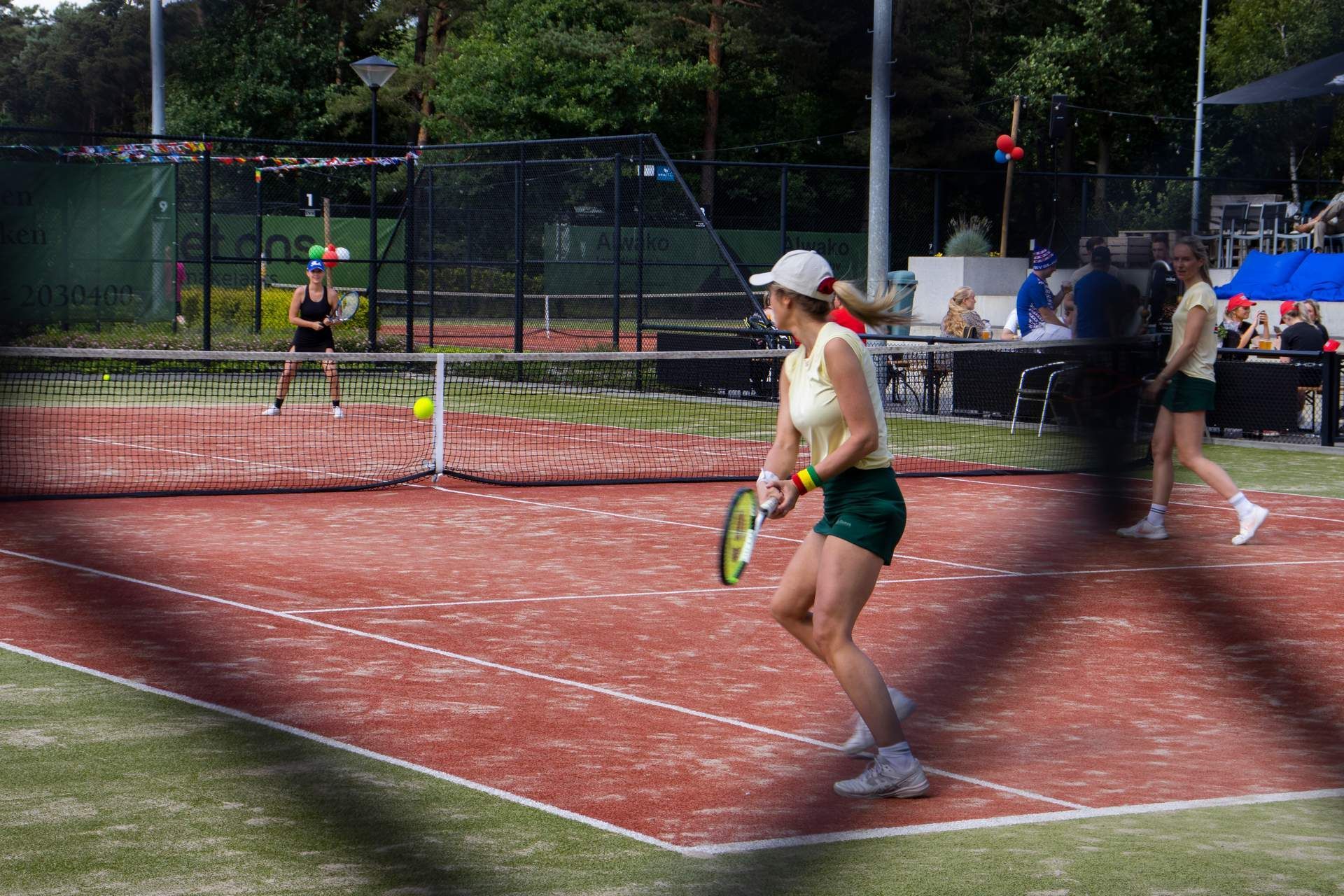 Mensen spelen padel op een gravelbaan met een net, een gele bal en in een buitenomgeving.