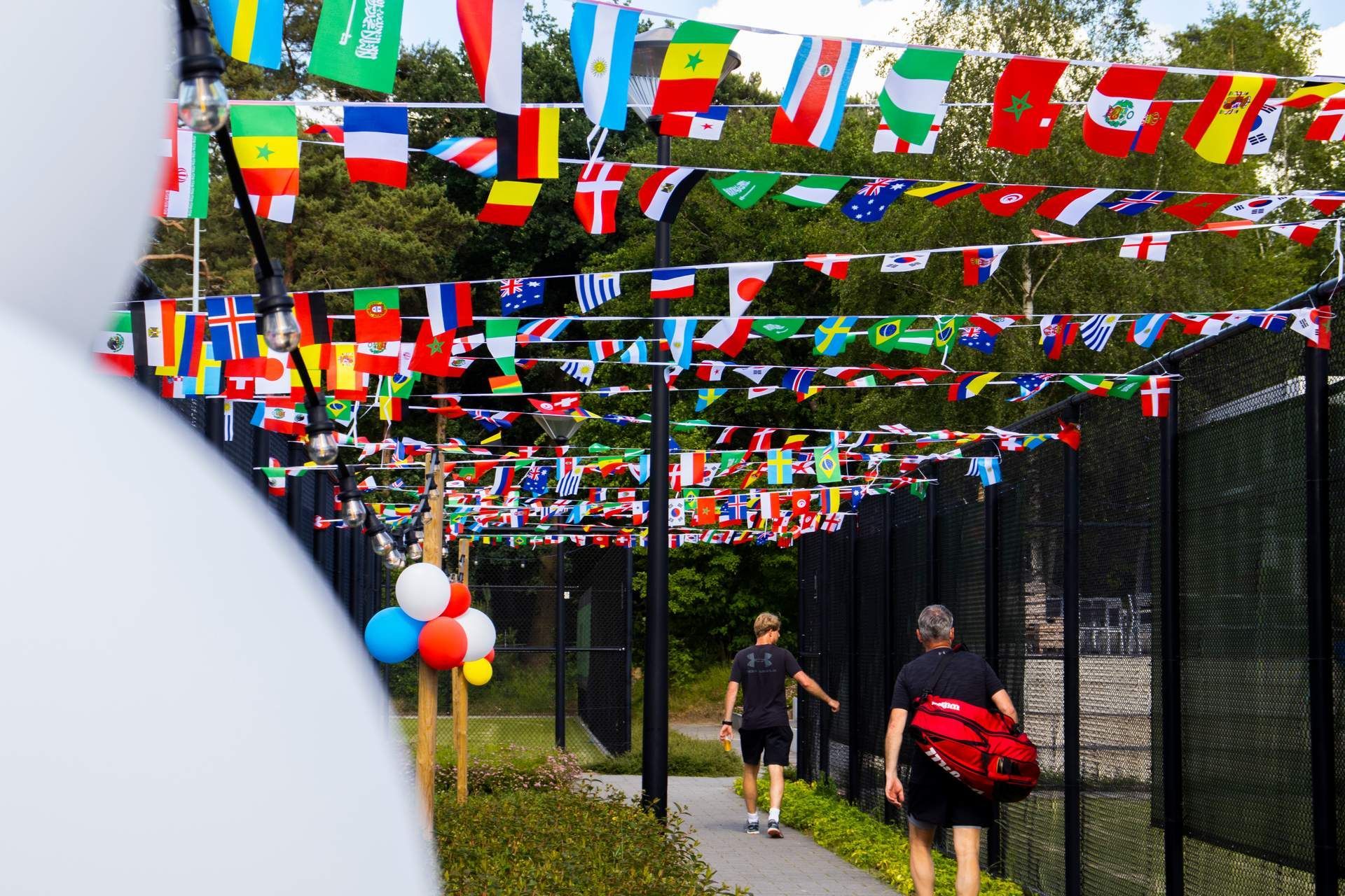 Mensen lopen over een pad onder rijen kleurrijke internationale vlaggen die boven hun hoofden hangen, vlakbij een tennisbaan.