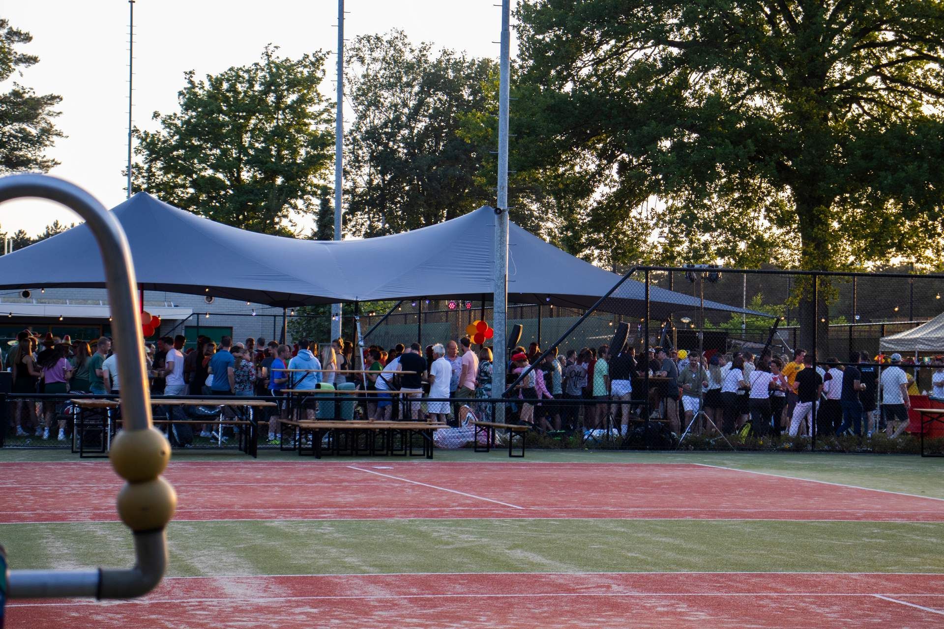 Een groep mensen praat gezellig met elkaar onder een groot, grijs afdak op een rood sportveld tijdens zonsondergang.