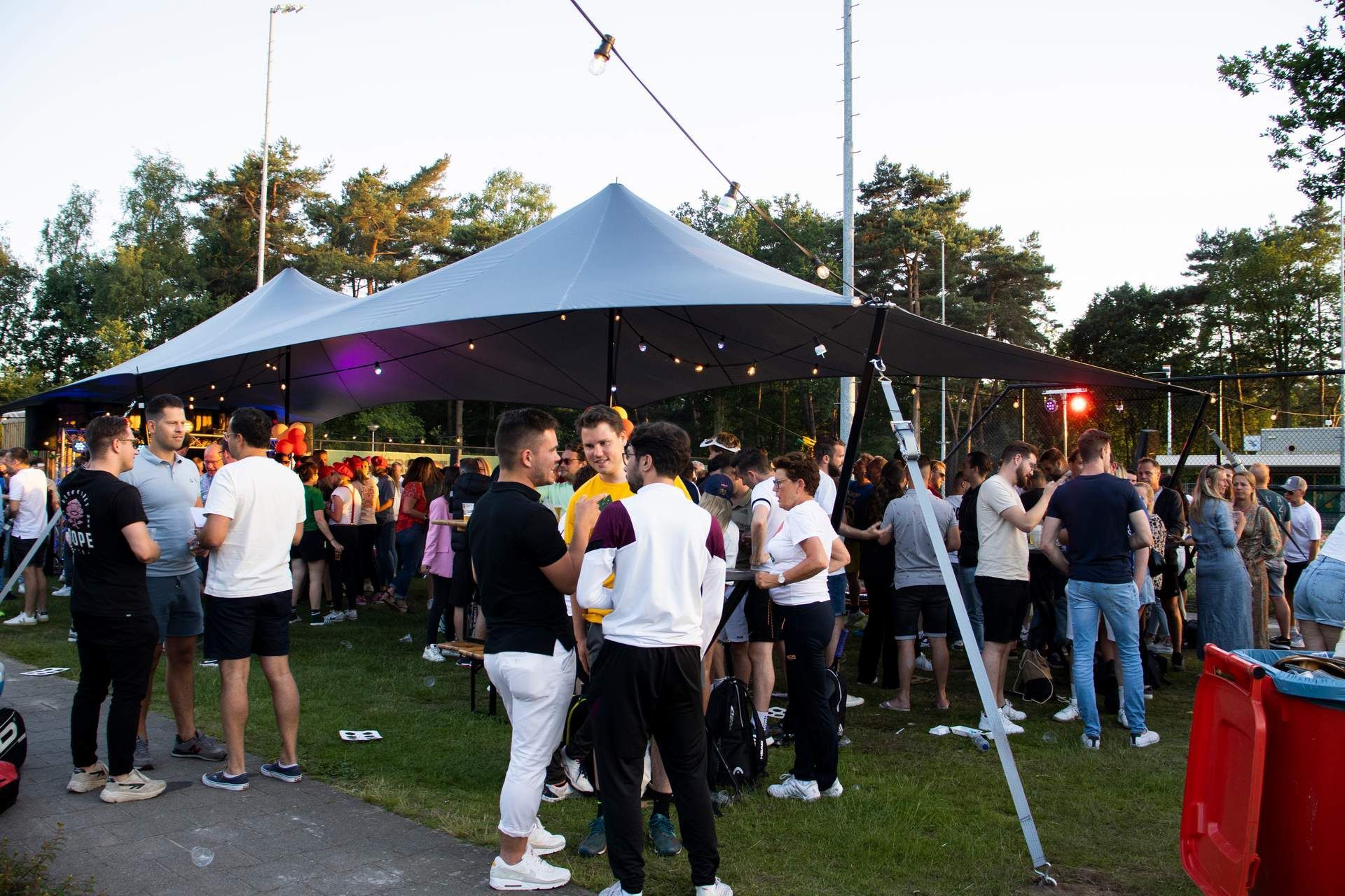 Mensen verzamelen zich onder een grote grijze partytent in een park tijdens een avondevenement.