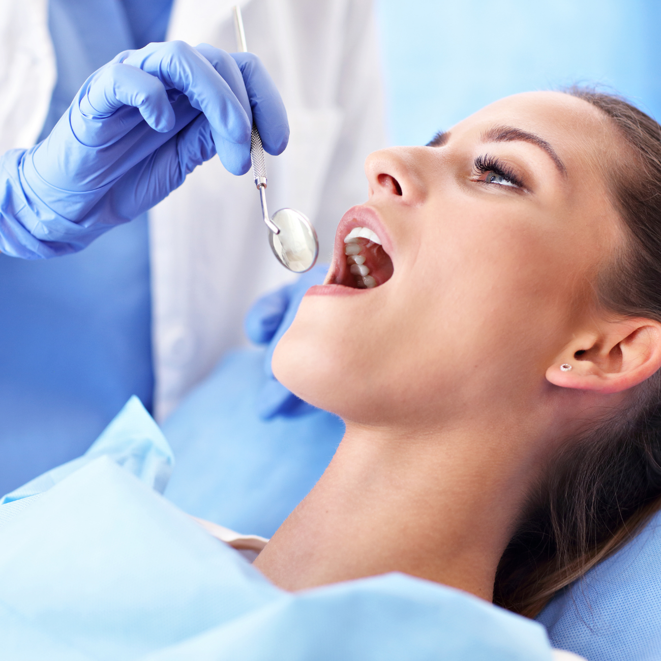Woman at dentist, mouth open, being examined with a mirror. Dentist wears blue gloves.