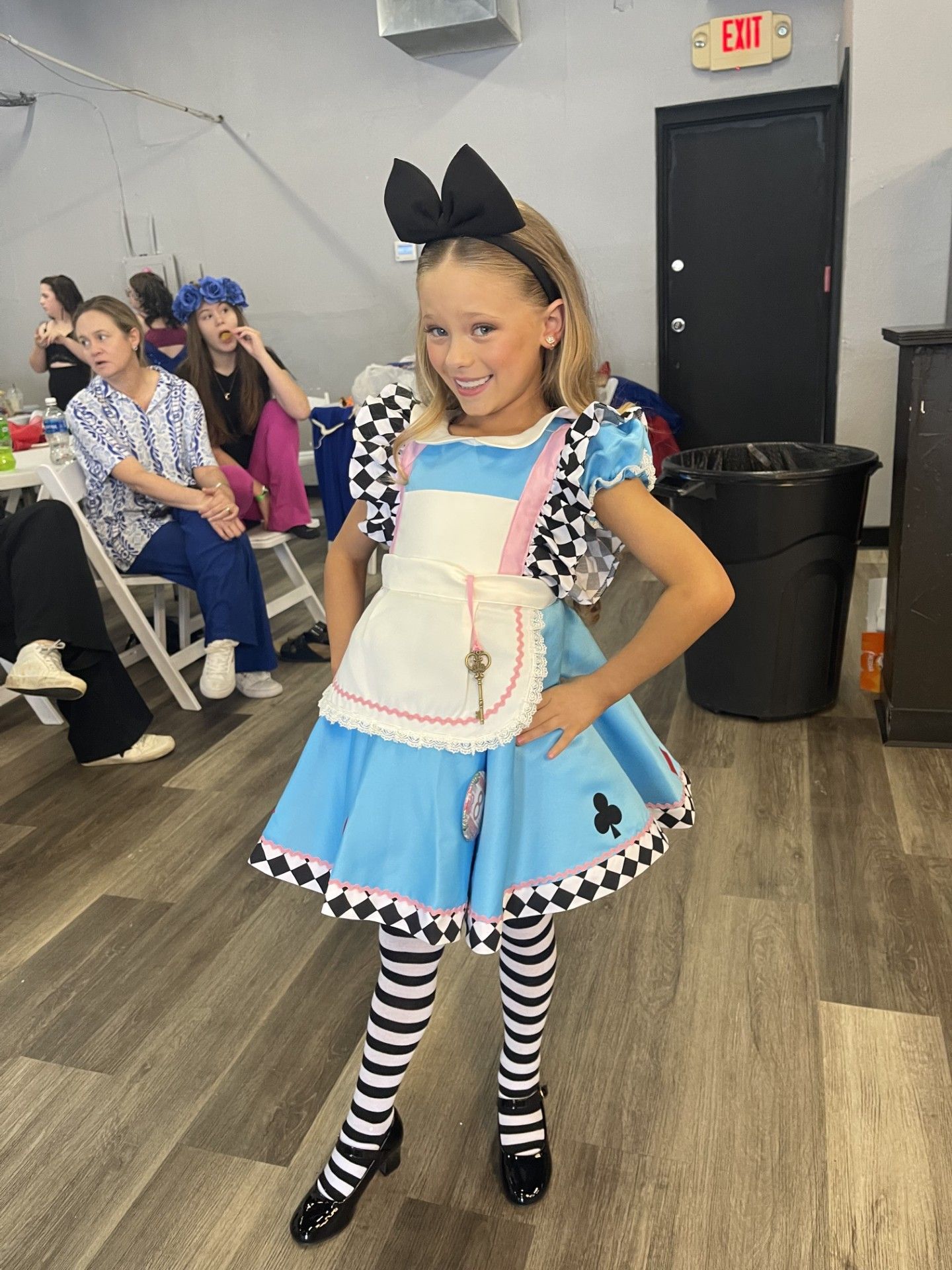Child in blue dress costume with striped tights and black bow, posing indoors at a party.