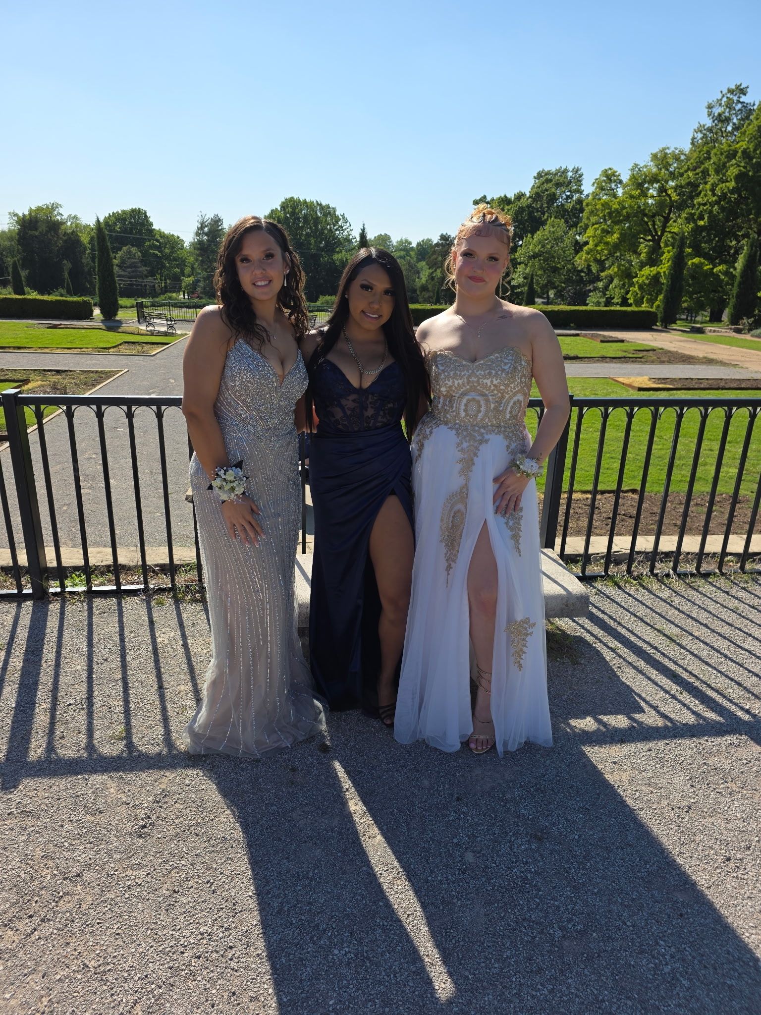 Three people posing outdoors in formal dresses by a black fence on a sunny day