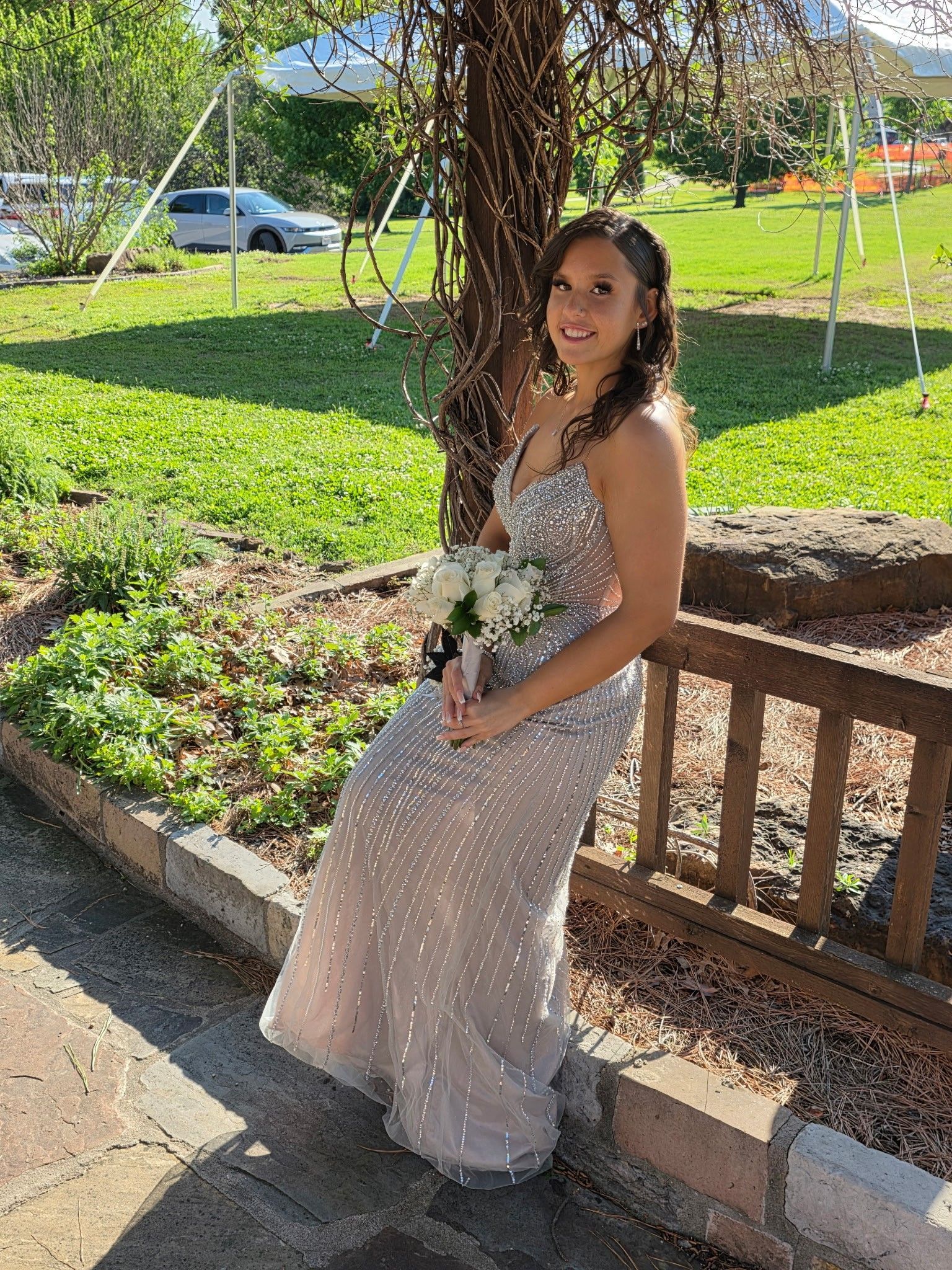 Woman in a sparkly dress sitting on a bench in a sunny garden, holding a bouquet and smiling.