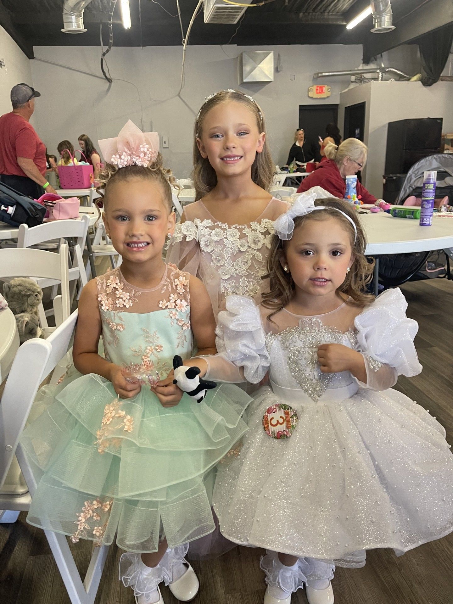 Three children in formal dresses holding small bouquets at a party table indoors