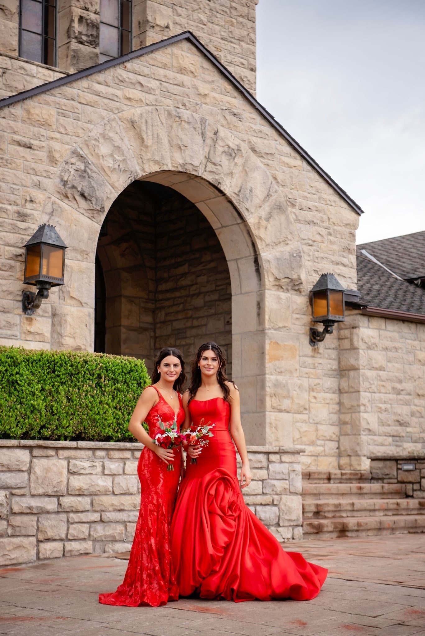 Two people in matching red gowns posing before a stone archway and wall