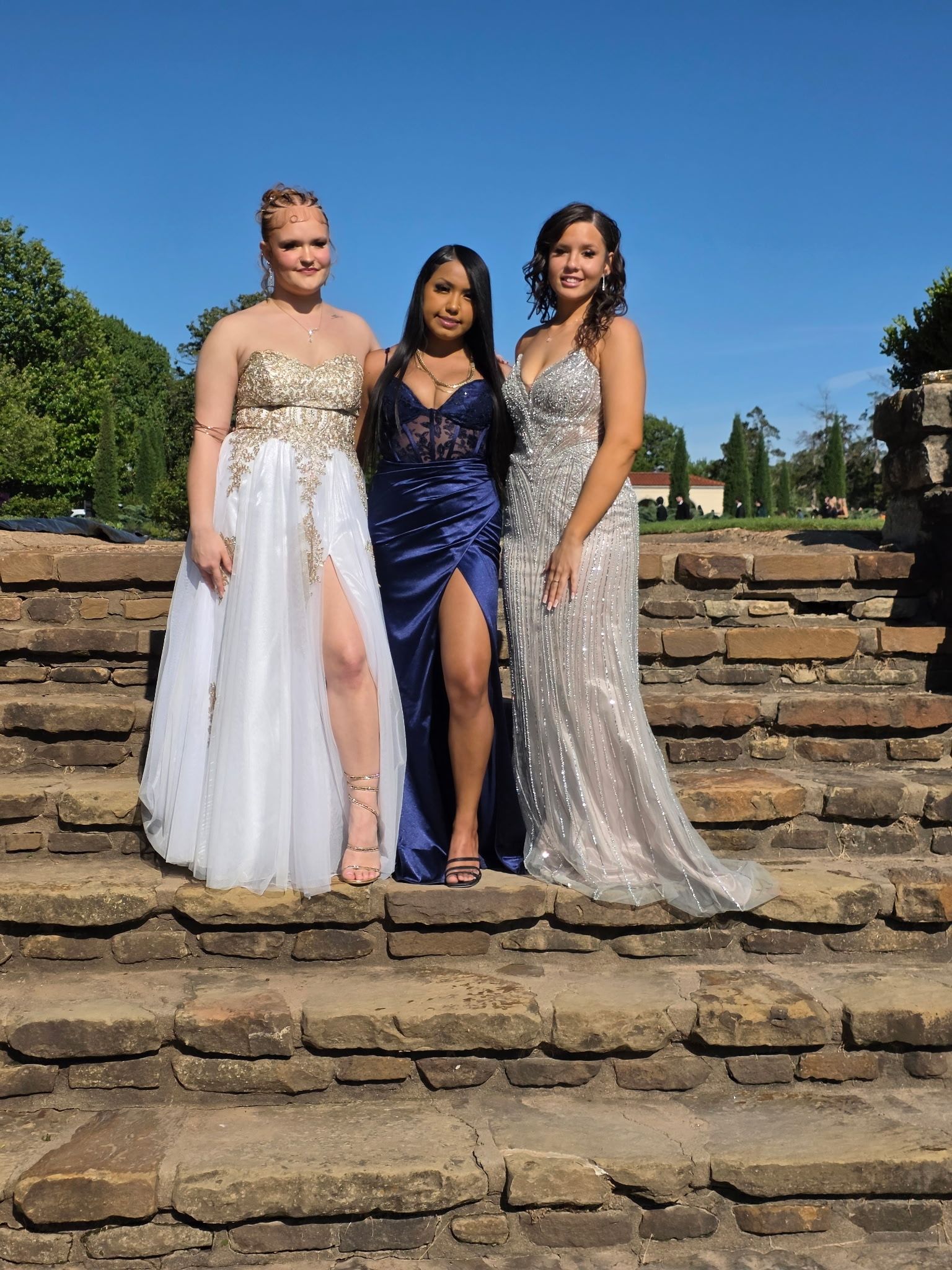Three people posing on stone steps outdoors in formal gowns under a clear blue sky.