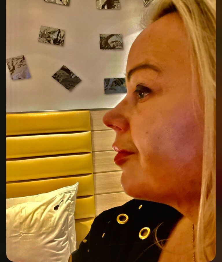 Woman with blonde hair, looking right, wearing a black shirt. Yellow headboard and wall with photos.