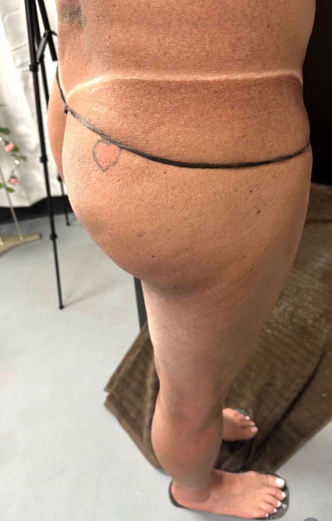 Person's lower body with tan lines, wearing a body chain with a heart, standing on a tiled floor.