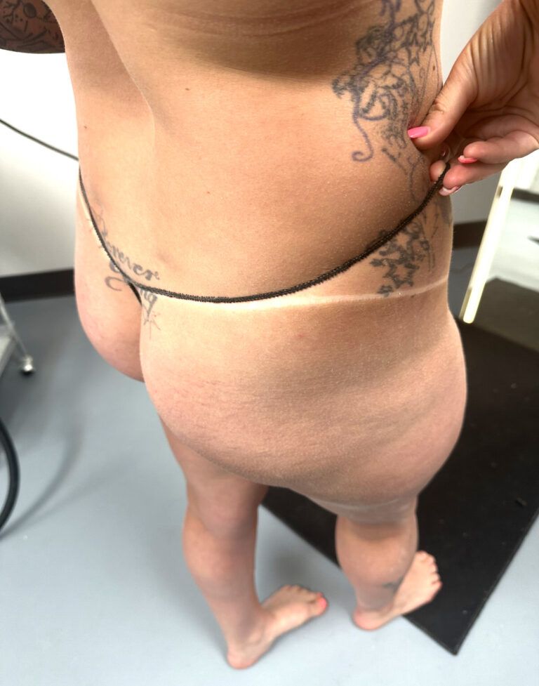 Person with a tan line, wearing a thong, with a tattoo on their back. Standing in a room with a black mat.