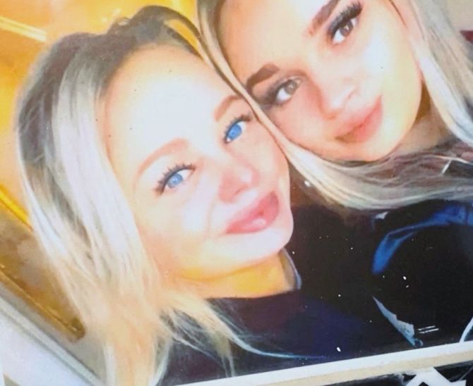 Two smiling people in a close selfie, one blonde and one brunette, against a warm indoor background