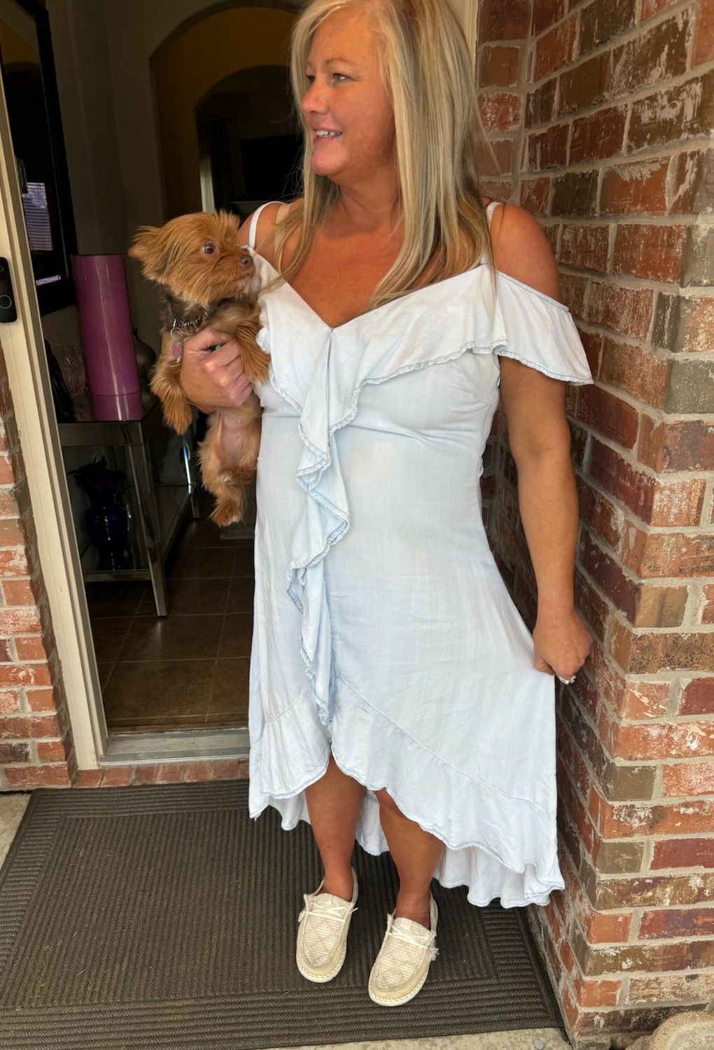 Woman in a white dress holding a small dog while standing by a brick wall at a doorway