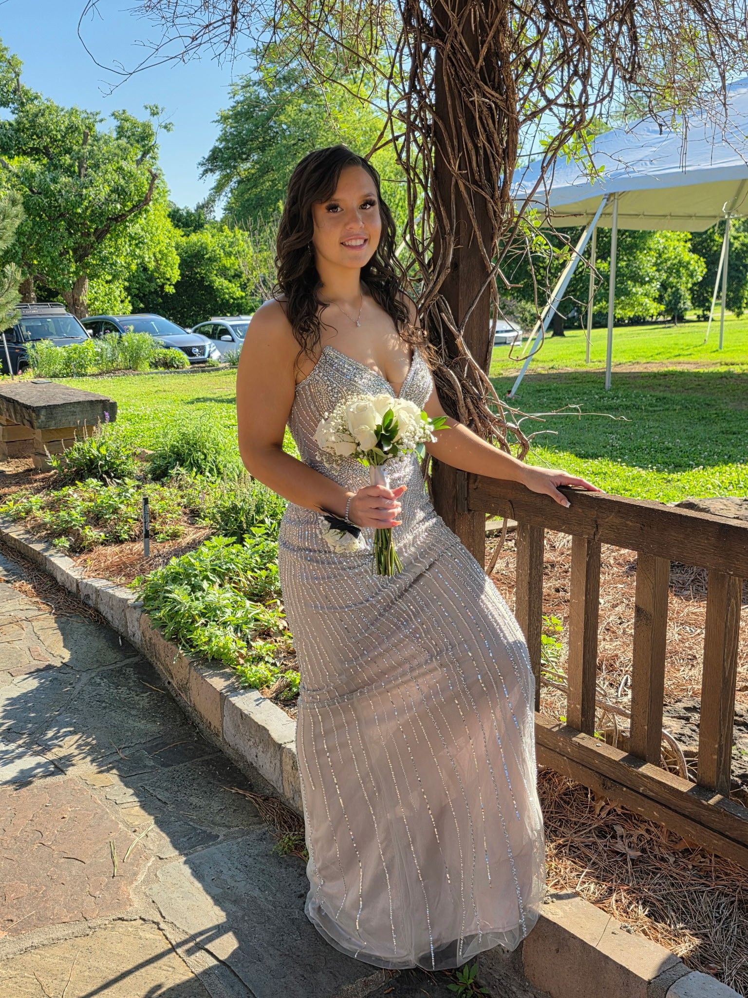 Woman in a white dress holding a bouquet by a wooden railing in a sunny garden.