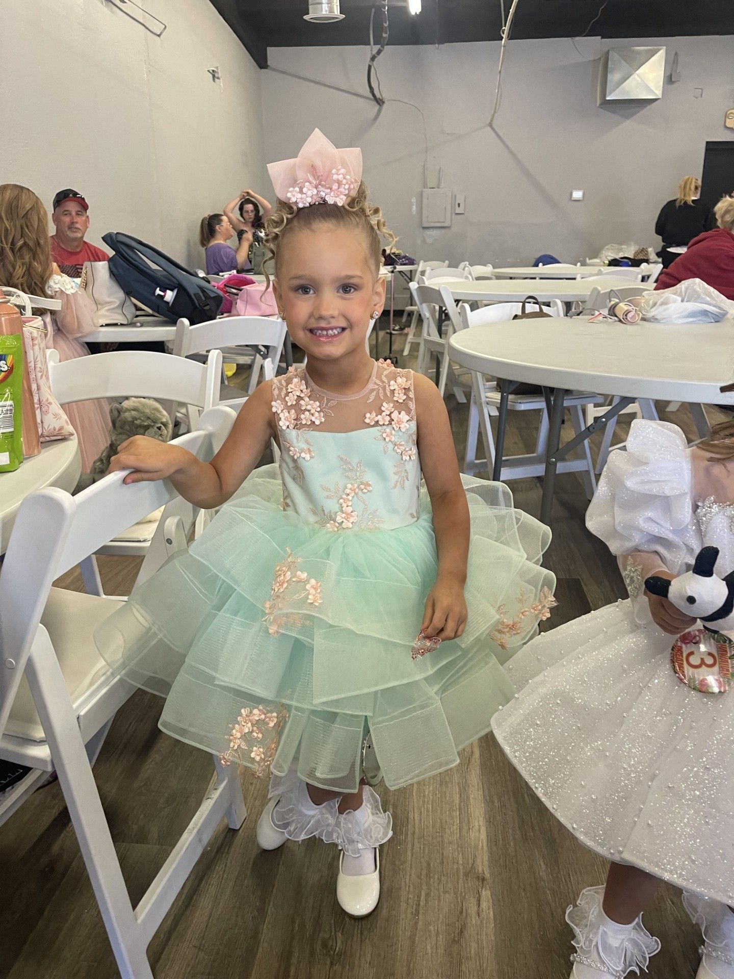 Young child in a mint green dress and large bow standing in a decorated event hall, smiling at the camera.