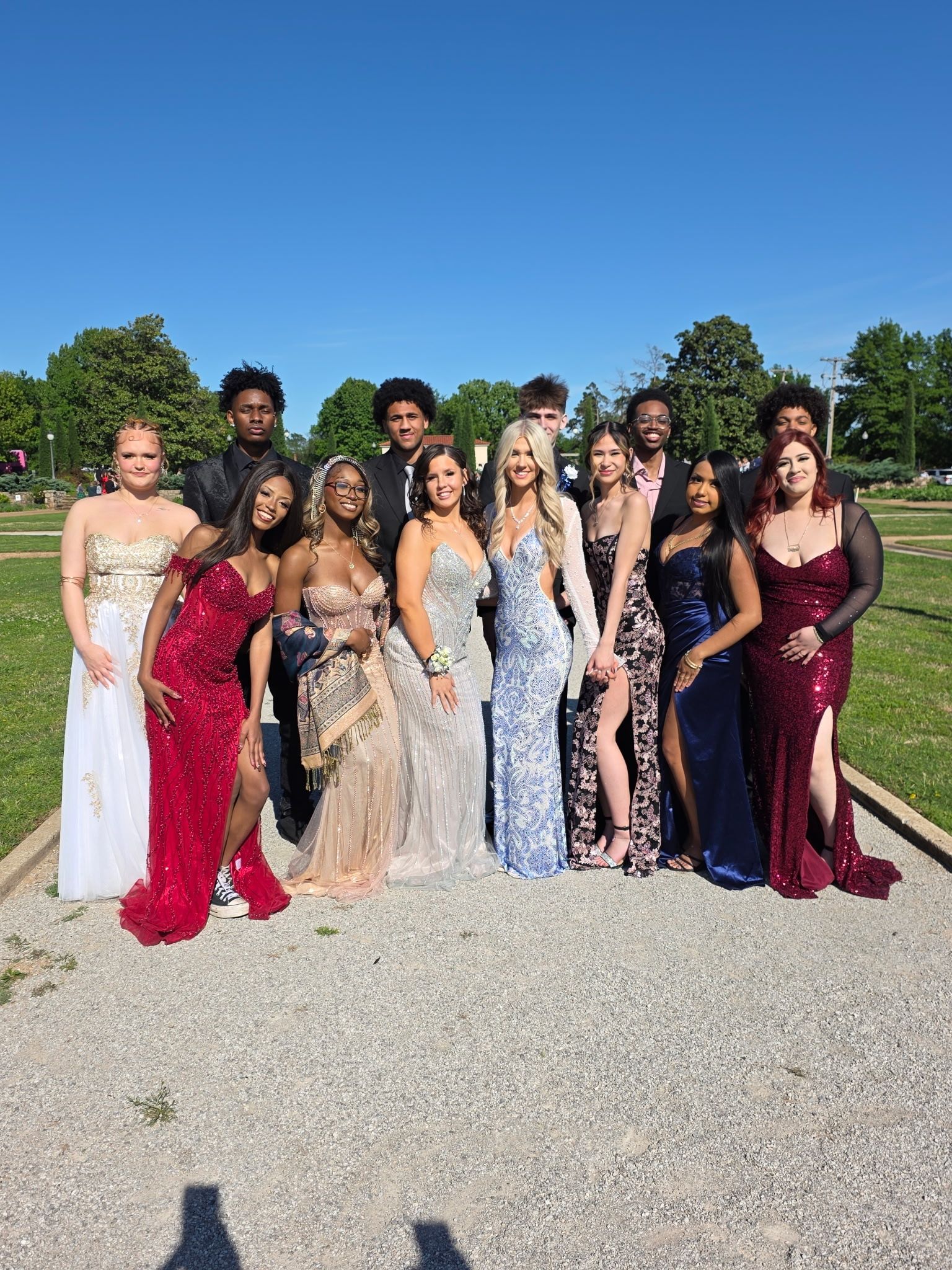 Group of people dressed in formal gowns and suits posing outdoors on a gravel path under a blue sky