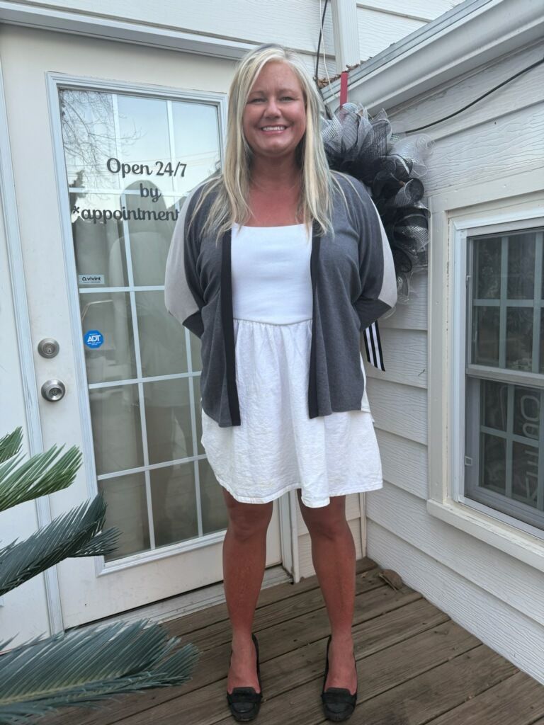 Woman standing on a porch in a white dress and gray cardigan, smiling in front of a glass door.