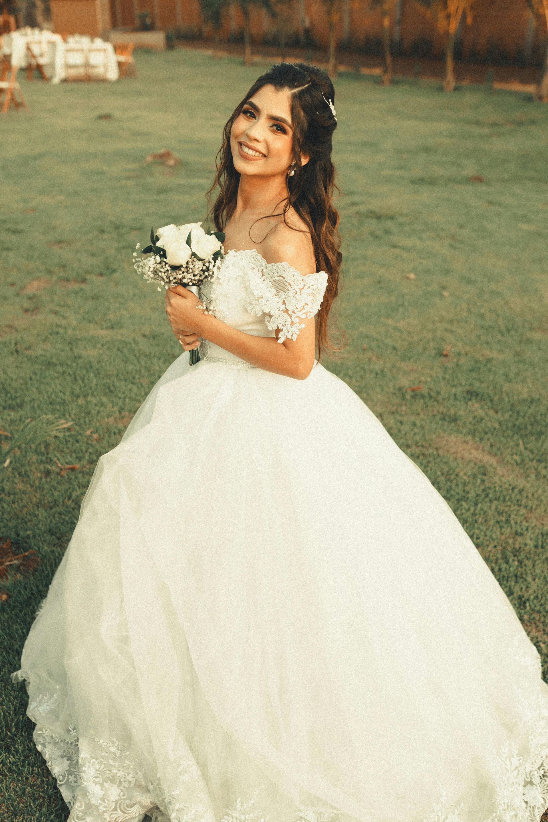 Bride in a white gown holding a bouquet, smiling in a grassy outdoor setting.