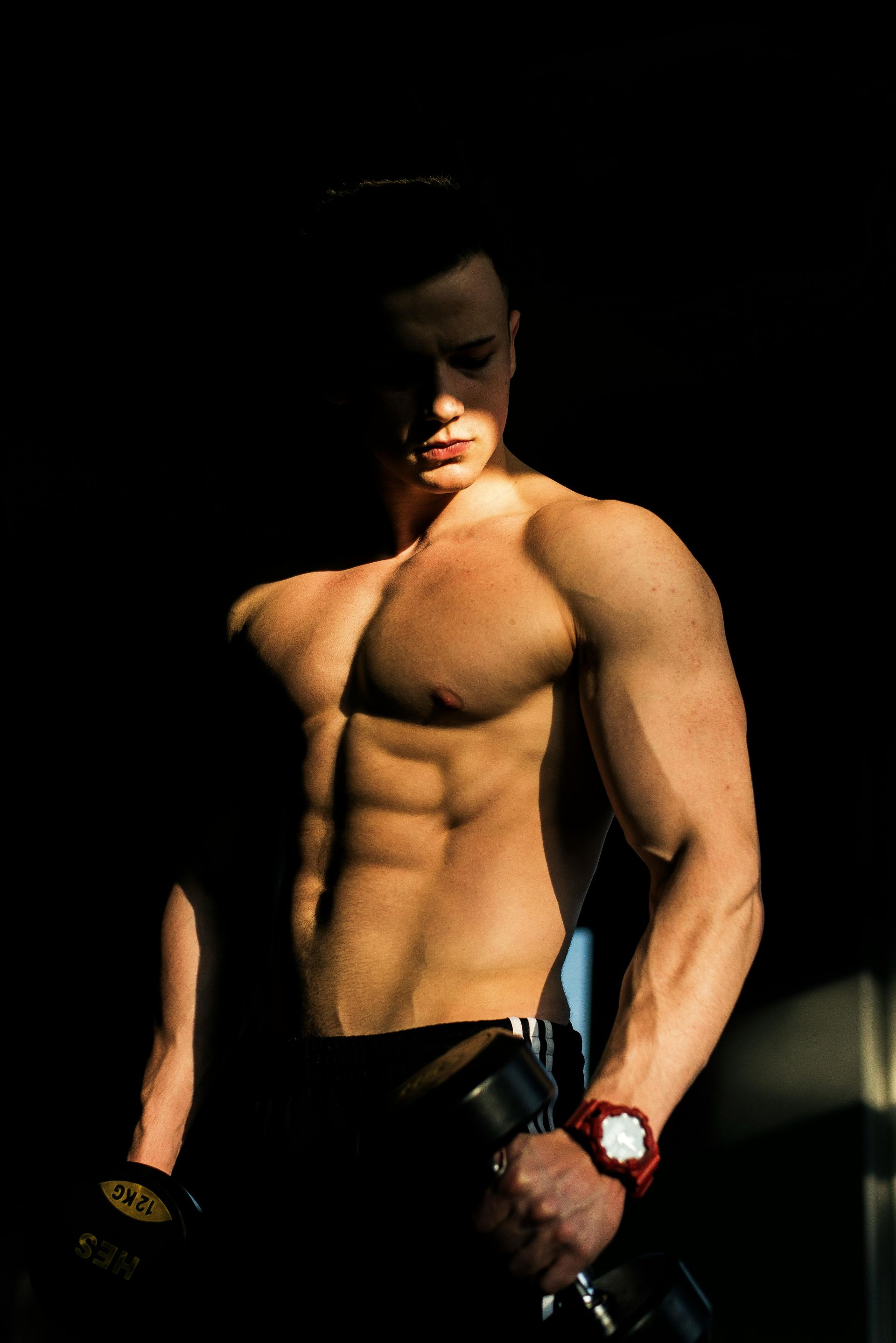 Shirtless man with defined muscles standing in dramatic low light, wearing dark pants and wristwatch.