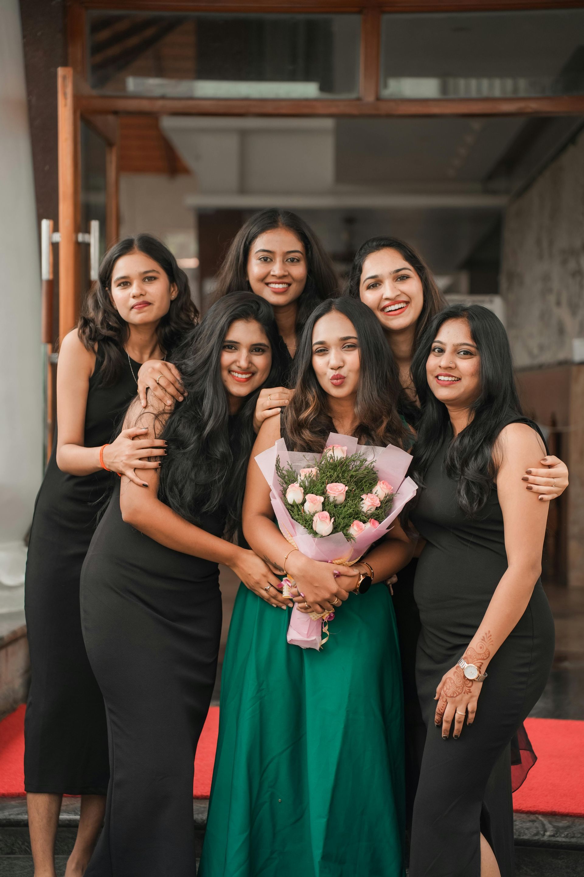 Five women in black and green formal dresses pose indoors; the center woman holds a bouquet.