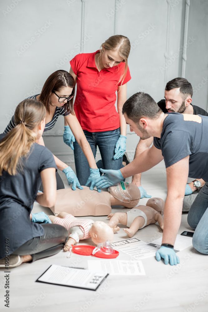 People practice CPR on a mannequin, with babies nearby, indoors.