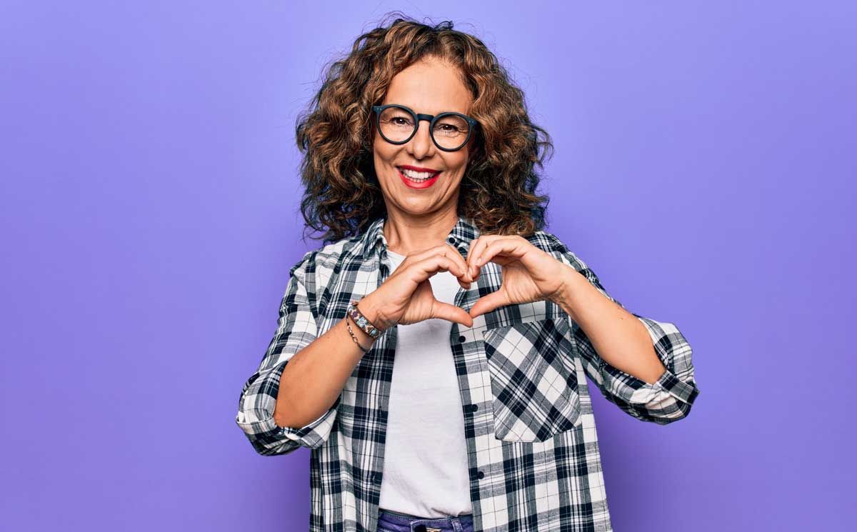 Woman with curly hair, glasses, and plaid shirt making heart with hands, smiling. Purple background.