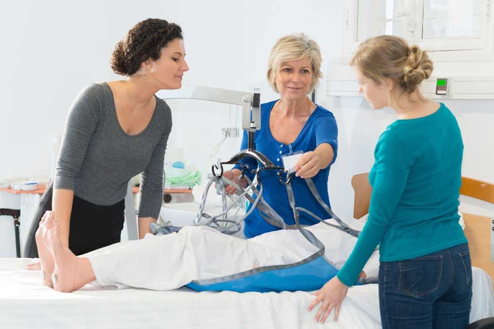 Three women demonstrating patient lift in a hospital room. The patient is under a white sheet.