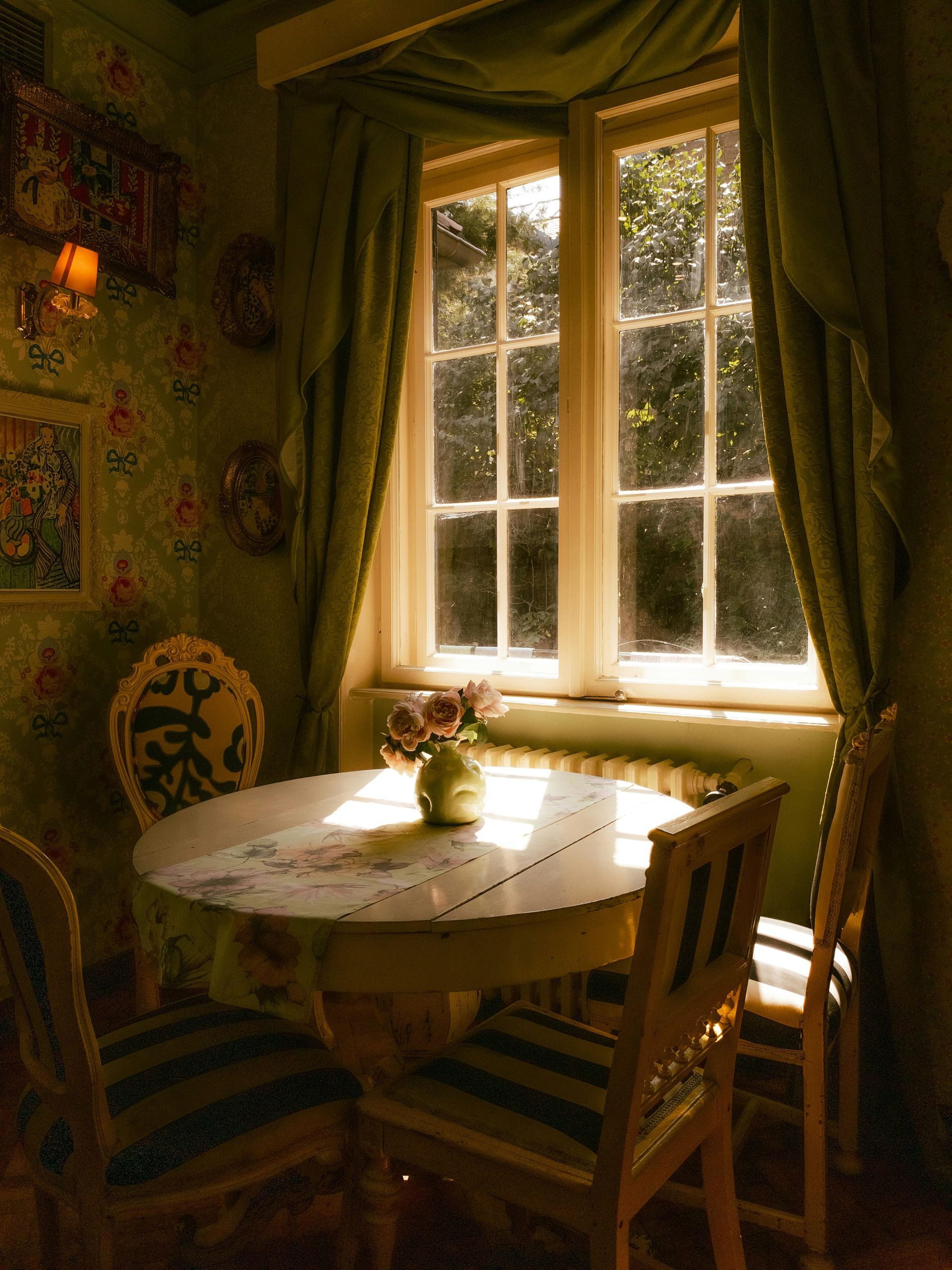 A cozy dining area with a round table and chairs, surrounded by floral wallpaper and a sunny window showcasing greenery outside.