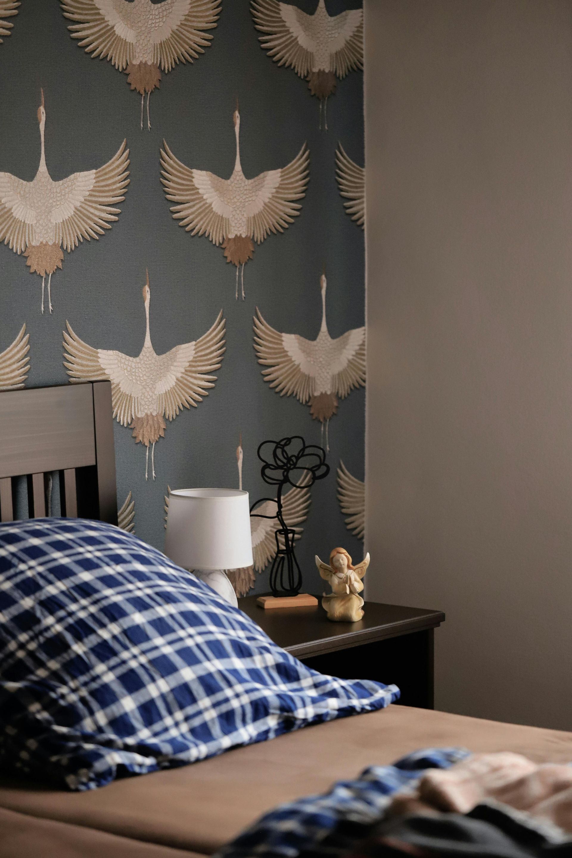 Bedroom with blue and white plaid pillow, patterned wallpaper with cranes, and a nightstand with a lamp.