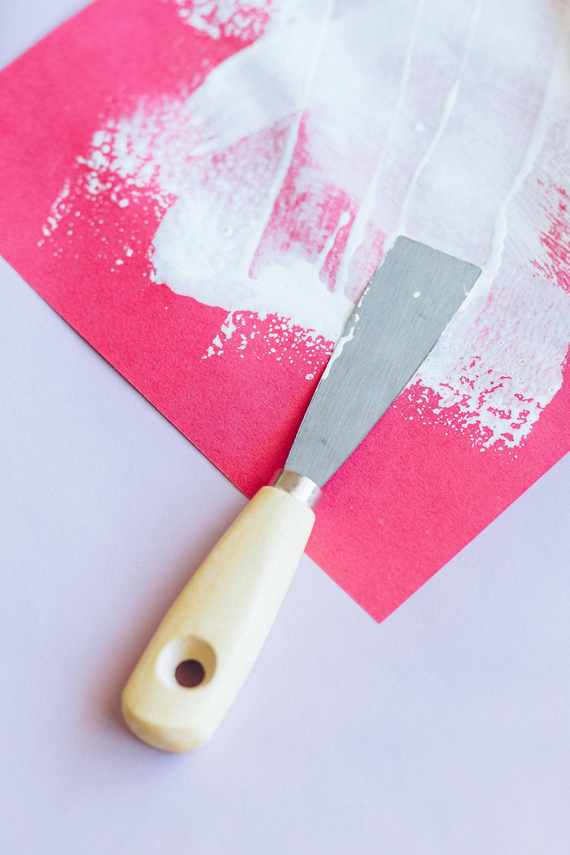 Close-up of a putty knife with a wooden handle spreading white paint across bright pink paper.