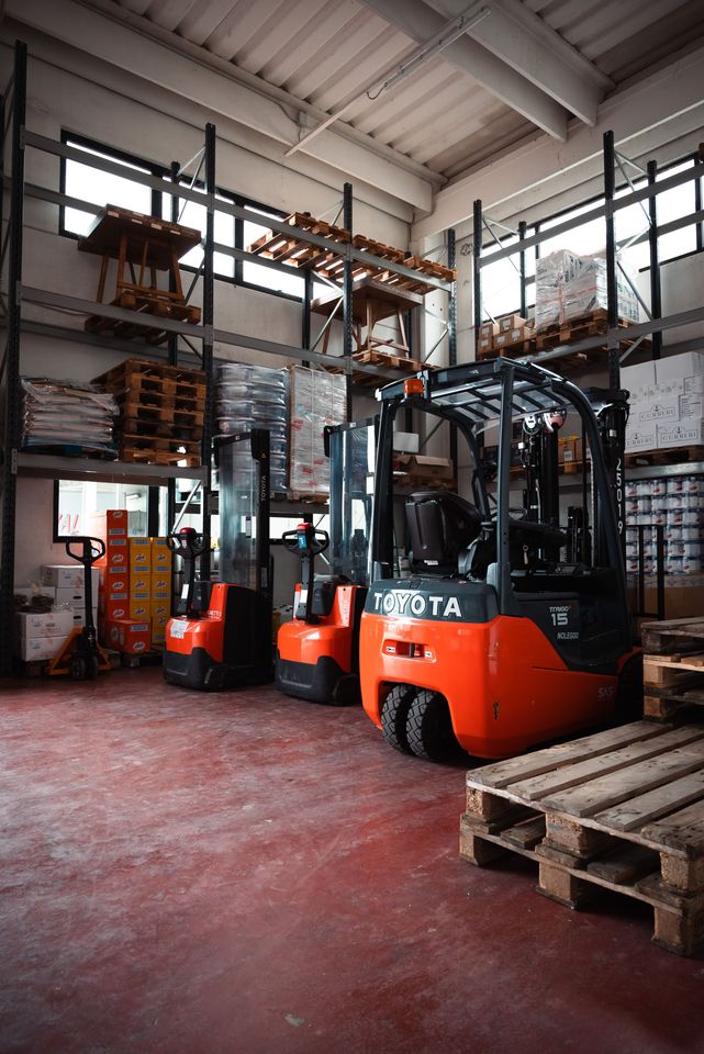 Tre carrelli elevatori Toyota arancioni parcheggiati all'interno di un magazzino con alte scaffalature metalliche e pallet di legno sul pavimento.